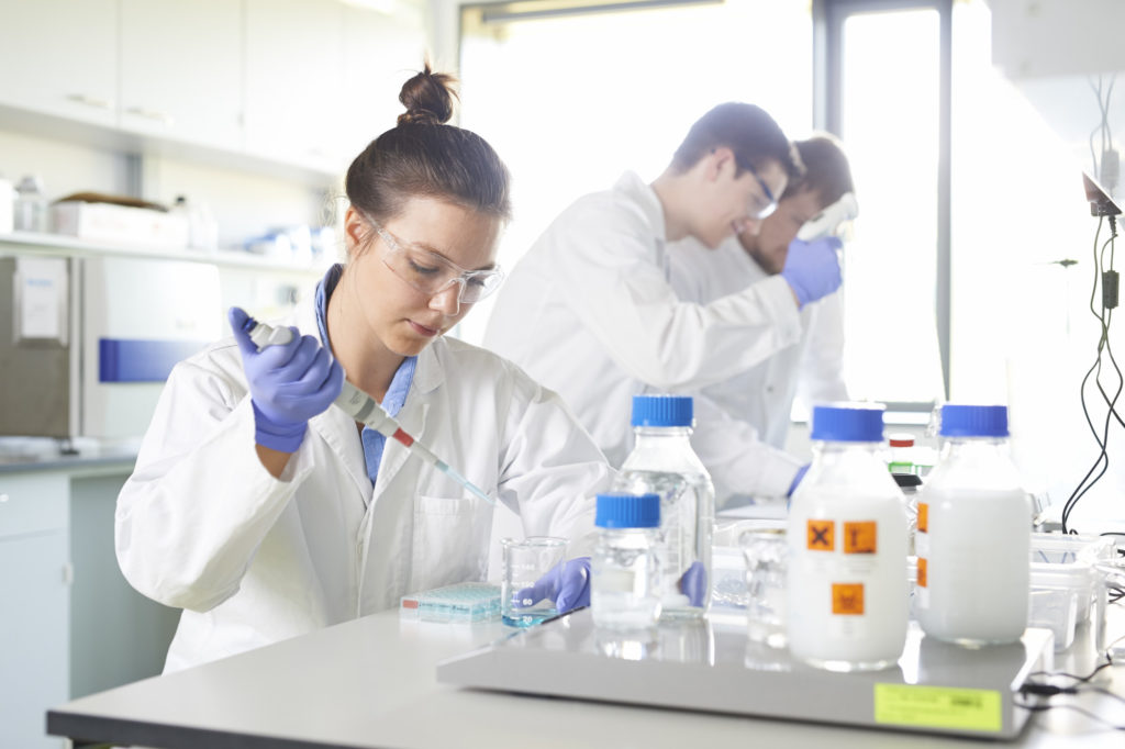 A young woman dressed in lab coat, gloves and specs pipettes a chemical substance in a laboratory.