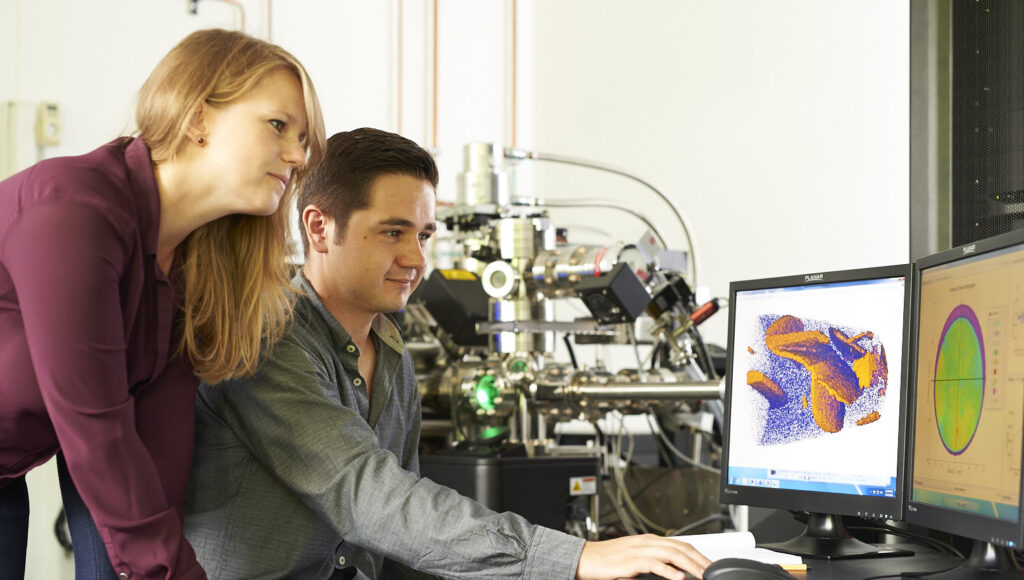 A young woman is standing next to a sitting man. Both are looking at two displays, where colored graphics are shown.