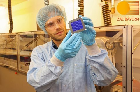 A member of the research team dressed in lab coat, specs and gloves is holding one of the new investigated organic solar modules. The solar module can be described as a metallic square plate. In the interior it’s blue with metallic vertical lines.
