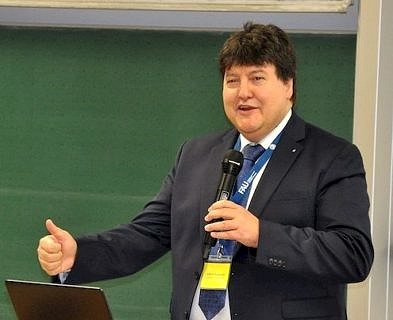 A man dressed in a dark suit with a blue tie and the blue FAU lanyard is holding a microphone. He’s giving/holding a speech regarding the 10th anniversary of the professorship WW7. He’s in a good mood while he’s giving the thumb.